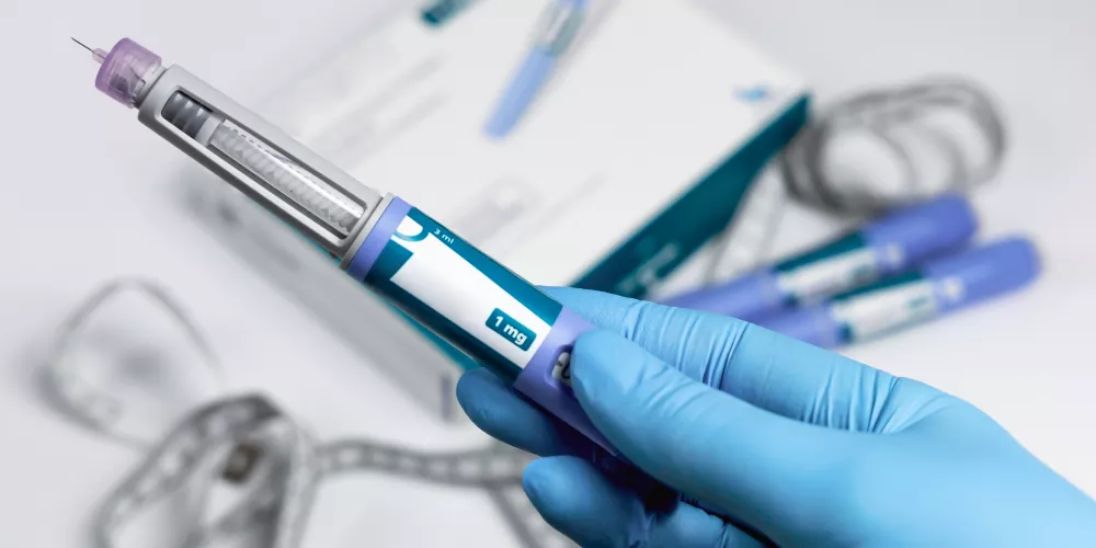 A gloved hand holds a blue injection pen with a visible needle. In the background, blurred boxes and similar pens suggest a medical setting.