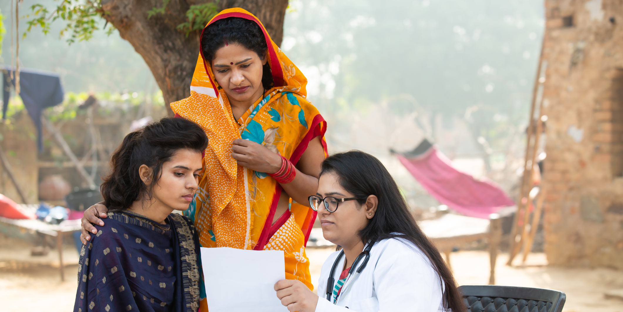 A doctor in a white coat examines a medical paper outdoors at a table with medicines. Beside her, a worried woman listens, with another woman standing supportively behind her. The setting is rural, with a hammock and tree in the background.